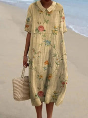 Abito estivo lungo floreale in lino, maniche corte, colletto a camicia, indossato su spiaggia. Borsa di paglia intrecciata. Moda donna casual.