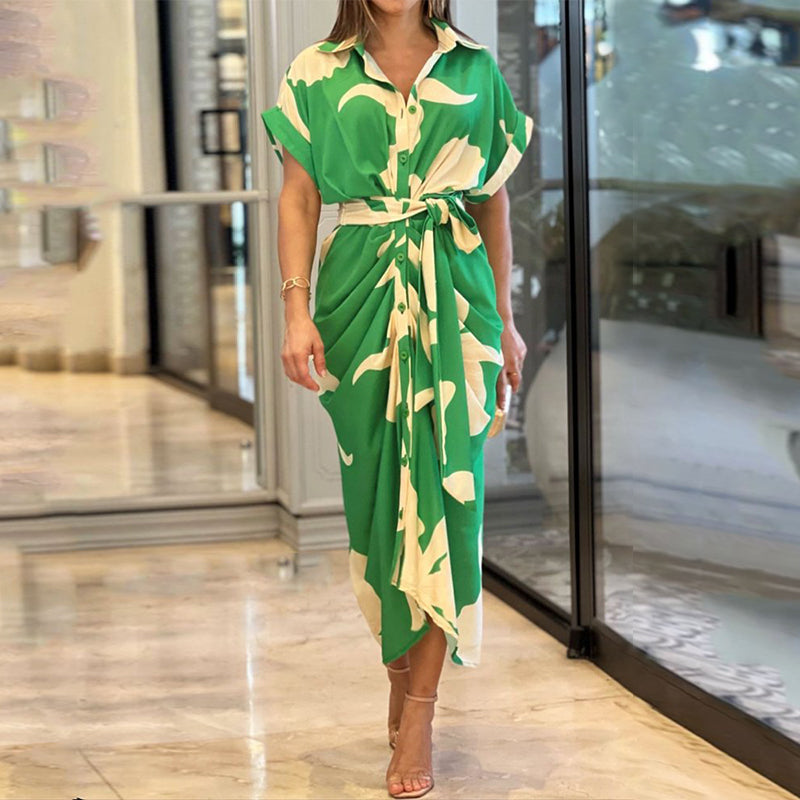 Abito lungo verde con stampa bianca, maniche corte e cintura in vita, indossato da una donna in un ambiente moderno. Moda estiva elegante.