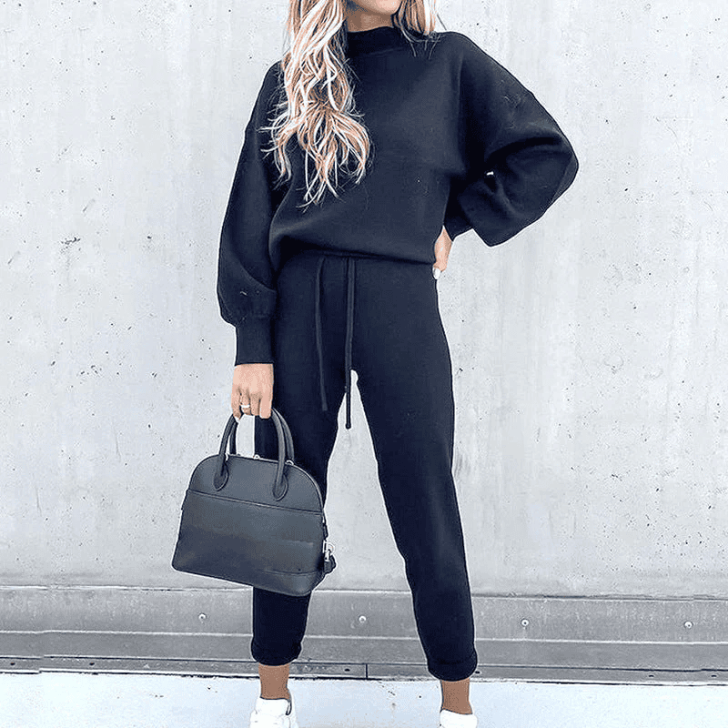 Donna con tuta nera casual, borsa a mano nera, sfondo grigio. Moda autunnale, abbigliamento comodo, stile urbano.