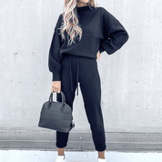 Donna con tuta nera casual, borsa a mano nera, sfondo grigio. Moda autunnale, abbigliamento comodo, stile urbano.