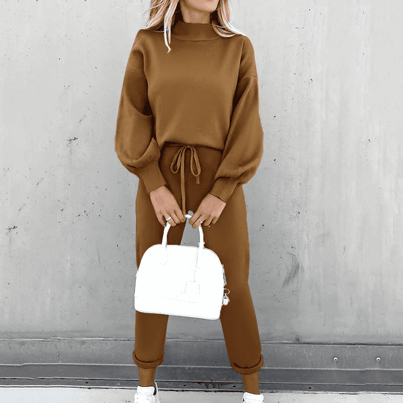 Donna con completo marrone a maglia, maglione a collo alto e pantaloni con coulisse, borsa bianca, scarpe da ginnastica bianche, sfondo grigio.