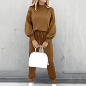Donna con completo marrone a maglia, maglione a collo alto e pantaloni con coulisse, borsa bianca, scarpe da ginnastica bianche, sfondo grigio.