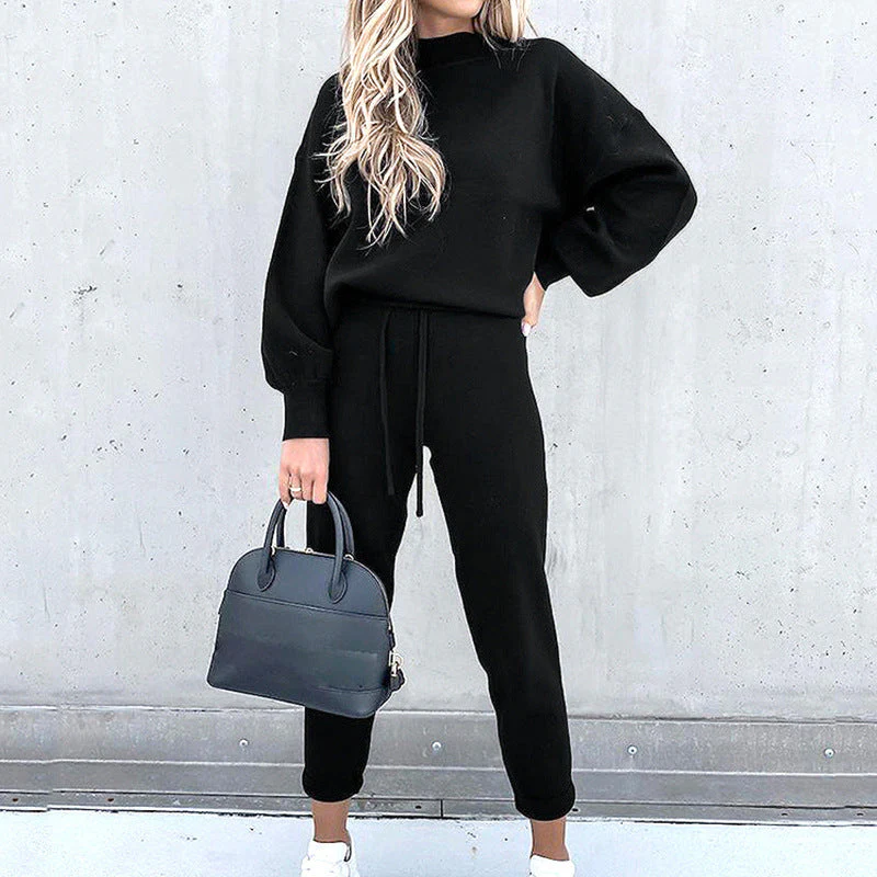 Donna con tuta nera elegante, borsa a mano blu scuro, sfondo grigio. Moda casual chic, abbigliamento autunnale, stile minimalista.