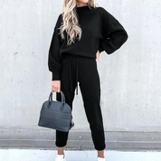 Donna con tuta nera elegante, borsa a mano blu scuro, sfondo grigio. Moda casual chic, abbigliamento autunnale, stile minimalista.